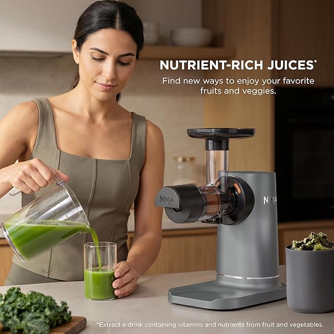 Cold press juicer with matte base and chute, with pitcher pouring green juice into a glass