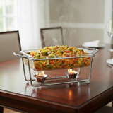 A glass rectangular chafing dish filled with a colorful pasta casserole sits on a metal stand with two small metal cups, placed on a polished wooden dining table with steam rising from the food.
